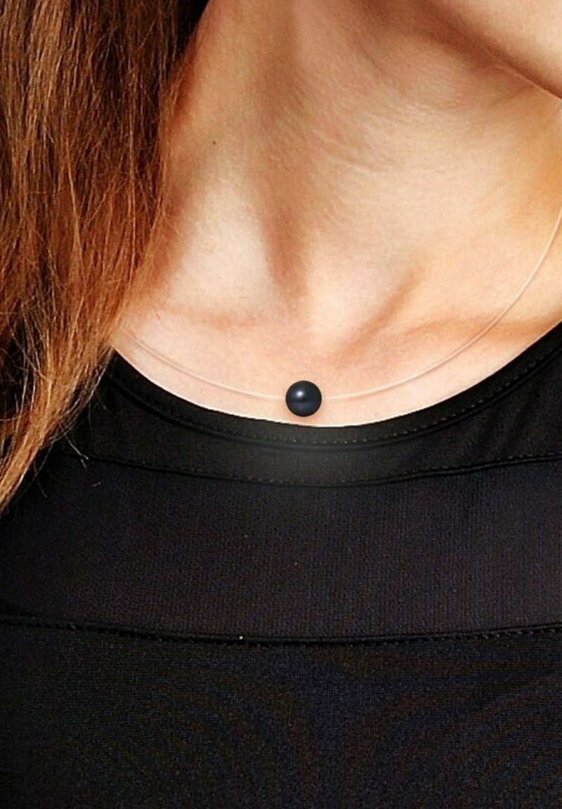 Collier pendentif clair avec une seule perle sphérique noire sur un cordon en nylon transparent fin, reposant sur un cou nu au-dessus d'un haut noir.