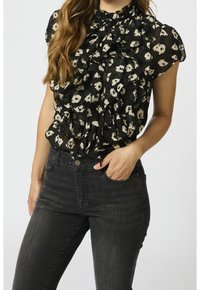 Svart genomskinlig blus med hög, rynkad krage och korta ärmar, med ett crèmefärgat blommönster. Matchas med mörka jeans.