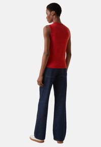 Czerwony bezrękawnik z dzianiny o dopasowanym kształcie, zestawiony z ciemnymi rozszerzanymi spodniami dżinsowymi z wysokim stanem oraz białymi trampkami. Prosty design.