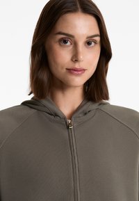 Olivgrüner Zip-Hoodie mit gerippter Textur und grauem Kapuzenfutter. Verfügt über einen Frontreißverschluss und ein körpernahes Design.