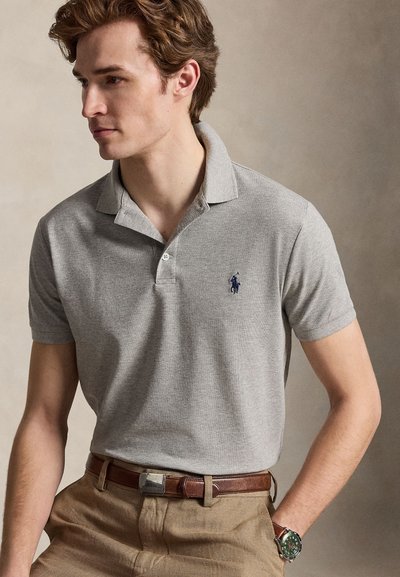 Jaunas vyras dėvi pilką polo marškinėlį, rudą diržą, šviesaus atspalvio kelnes ir žalią laikrodį ant riešo, žiūri į kairę, neutralioje fone.