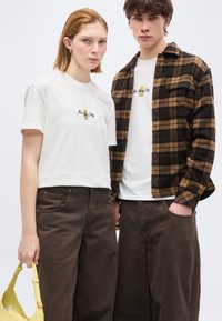 Νεαρή γυναίκα και άντρας φορούν λευκά μπλουζάκια με γραφικά και καφέ παντελόνια, με τον άντρα να φορά καφέ καρό πουκάμισο και τη γυναίκα να κρατά κίτρινη τσάντα.