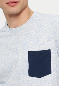 Κοντινό ενός ατόμου που φοράει ένα ανοιχτό μπλε μπλουζάκι με σκούρη μπλε τσέπη στο στήθος.