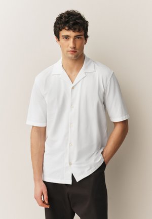 Mladý muž s kudrnatými vlasy v krátkém bílém košili s knoflíky a černých kalhotách, stojící před jednoduchým světlým pozadím.