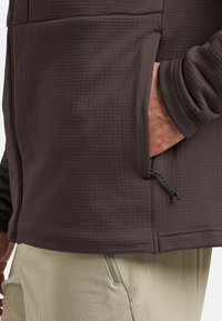 Giacca marrone scuro, testurizzata, con tasca laterale e cordino regolabile. Indossata sopra pantaloni chiari con tasca con zip.