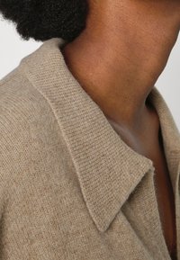 Beige stickad tröja med bred krage. Mjuk textur och ett fint detaljert mönster i varierande nyanser av brunt.