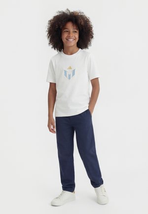 Mosolygó, göndör hajú gyermek fehér Adidas logós pólót, tengerészkék nadrágot és fehér tornacipőt viselve áll semleges háttér előtt.