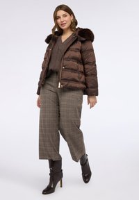Hnědá péřová bunda s kapucí podšitou kožešinou, kombinovaná se vzorovanými kalhotami s vysokým pasem. Modelka má na sobě hnědé kozačky na vyšším podpatku až po kolena.
