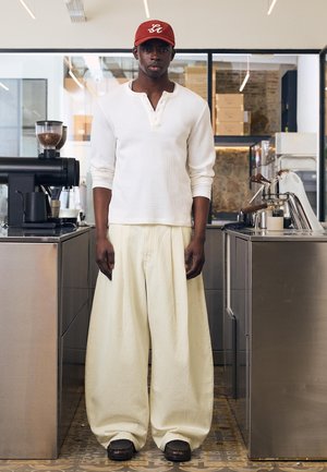 Mladić u crvenoj kapi, bijeloj majici dugih rukava i širokim krem hlačama stoji između nehrđajućih čeličnih aparata za kavu u kafiću.