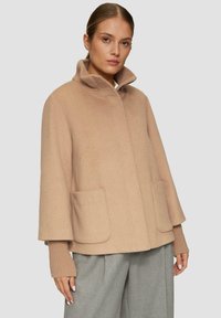 Beige Wollmantel mit hochgeschlossenem Kragen, seitlichen Taschen und gestrickten Ärmelakzenten. Das Design zeichnet sich durch eine lockere Passform und einen strukturierten Stoff aus.