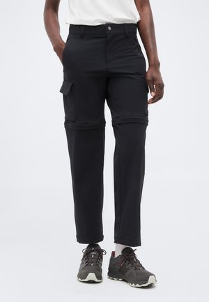 Pantaloni cargo negri cu picioare detașabile, purtați cu pantofi gri de drumeție și șosete albe, pe o persoană cu mâinile în buzunare.