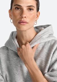 Šedá mikina s kapucňou a šnúrkou, s jemnou textúrou a jemnými ozdobami. Model drží látku v ruke, na ktorej sú prirodzené nechty.