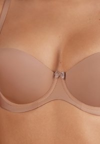 Beige gevoerde beha met een gladde oppervlakte, onderwired ondersteuning en decoratieve metalen strikdetails in het midden tussen de cups.
