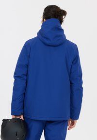 Blå skidjacka med huva, tillverkad av ett slätt, vattentätt material. Har rak passform och kombineras med en svart hjälm.