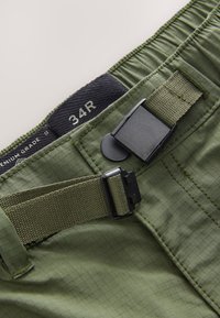 Pantaloncini verde oliva con motivo a griglia texturizzata, dotati di fibbia nera regolabile e etichetta intrecciata sulla vita con la scritta '34R'.