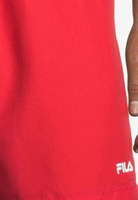 Röd sportt-shirt gjord av ett glatt och lätt tyg. Har en vit Fila-logotyp i det nedre högra hörnet. Enkel design utan mönster.