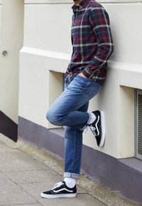 Koszula w kratę zapinana na guziki w kolorach granatowym, czerwonym i kremowym; jasnoniebieskie dżinsy; czarno-białe trampki z białymi skarpetkami; stojąca przy ścianie.