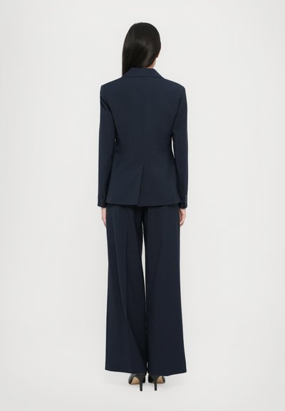 Completo blu navy su misura con giacca strutturata e pantaloni a gamba larga. Tessuto liscio e linee pulite senza motivi o decorazioni visibili.
