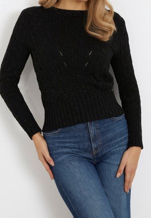 Pull en maille côtelée noir à manches longues, avec un motif texturé et de petites découpes à l'avant, associé à un jean bleu.