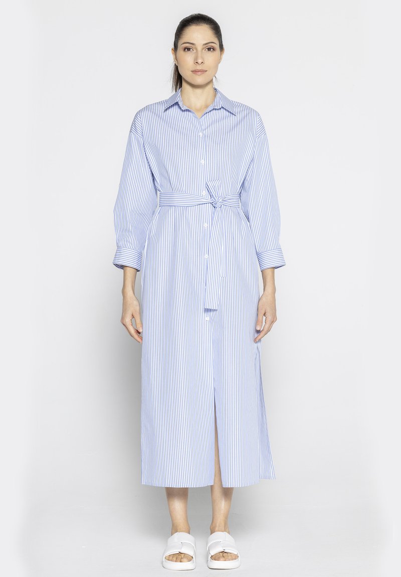 Niebieska i biała w paski długa sukienka koszulowa z długimi rękawami, guzikami z przodu, przewiązana w talii i z bocznym rozcięciem. W zestawie z białymi sandałami.