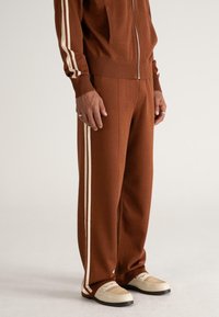 Persoană îmbrăcată în pantaloni de trening maro și jachetă cu fermoar asortată, cu dungi albe pe margini, purtată cu loaferi din pânză bej.