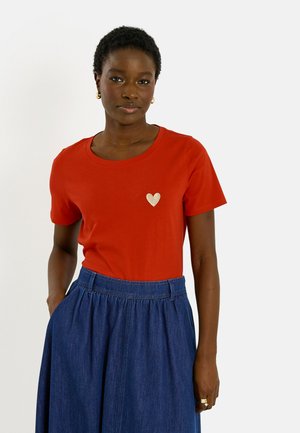 Femme portant un T-shirt rouge avec un petit cœur beige sur la poitrine et une jupe en jean bleue taille haute, debout avec une main dans la poche.