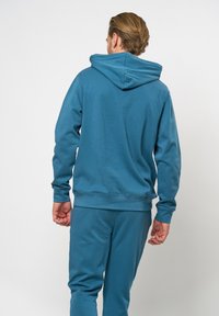 Kapuce mikina v barvě teal, s dlouhými rukávy a uvolněným střihem. Hladký materiál s jednoduchým designem, zobrazená zadečku. Odpovídající tepláky.