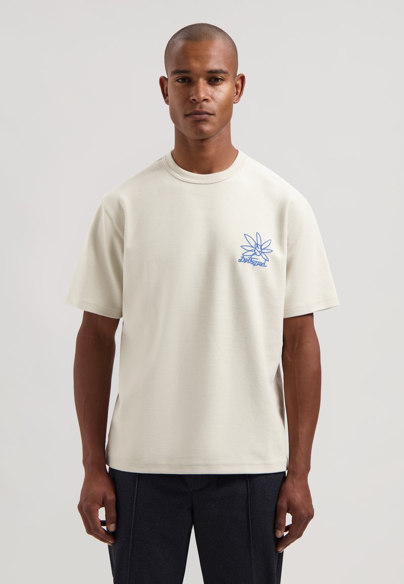 Camiseta de manga corta de color beige claro hecha de tela texturizada, con un diseño bordado azul en el pecho izquierdo. Cuello redondo sencillo.