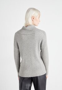 Pull en maille côtelée gris clair avec un col haut et des manches longues ; présente une silhouette ajustée et une texture douce. Un pantalon sombre est visible en dessous.