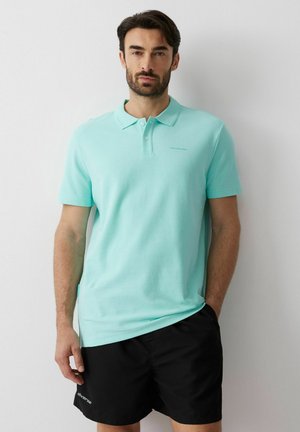 Mężczyzna w jasnoniebieskiej koszulce polo i czarnych szortach stoi przy białej ścianie z jedną ręką w kieszeni.