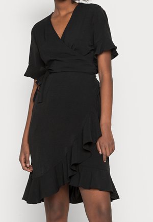 Kobieta ubrana w czarną, krótkim rękawem sukienkę kopertową z falbankami na krawędziach i wiązaniem w pasie, stojąca na tle jasnego, jednolitego tła.