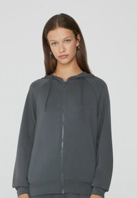 Sudadera con cremallera gris, de textura suave, con mangas raglán y cordones ajustables en la capucha. Cuenta con una cremallera frontal y puños acanalados.