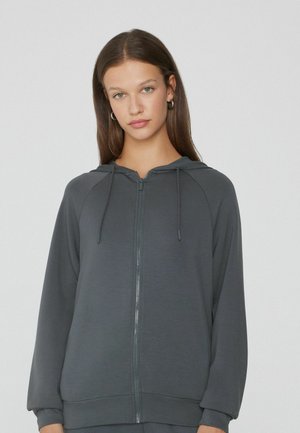 Sweat à capuche zippé gris avec une texture lisse, manches raglan et cordons de serrage réglables au niveau de la capuche. Comprend une fermeture éclair sur le devant et des poignets côtelés.