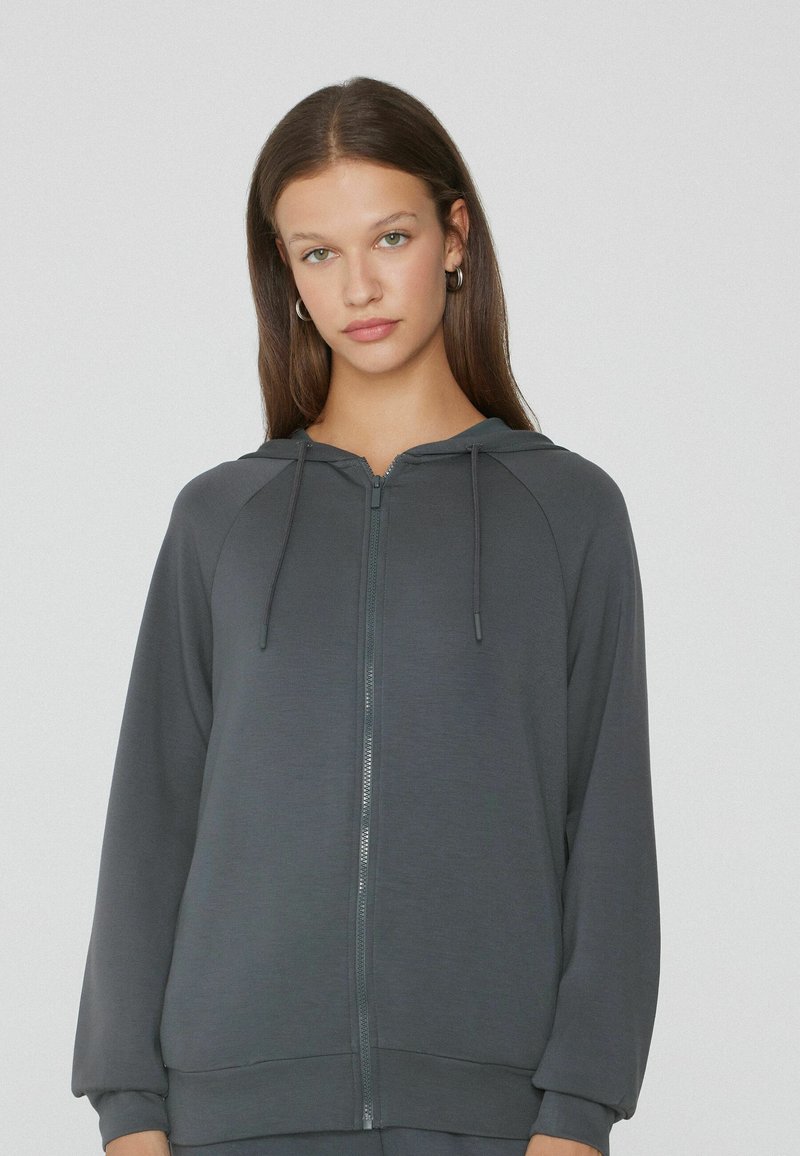 Sweat à capuche zippé gris avec une texture lisse, manches raglan et cordons de serrage réglables au niveau de la capuche. Comprend une fermeture éclair sur le devant et des poignets côtelés.