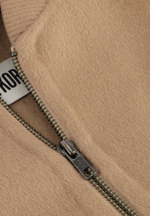 Beige Fleecejacke mit einem Metallreißverschluss; besitzt gerippte Bündchen und eine weiche Textur; Etikett mit Markennamen an der inneren Naht sichtbar.
