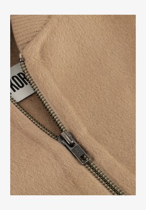 Beige Fleecejacke mit einem Metallreißverschluss; besitzt gerippte Bündchen und eine weiche Textur; Etikett mit Markennamen an der inneren Naht sichtbar.