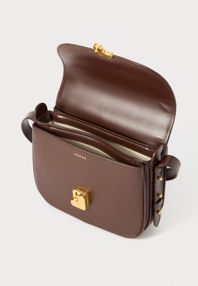 Brūna ādas crossbody soma ar zelta sprādzi, regulējamu siksnu un atvērtu augšu, kas atklāj gaiši brūnus iekšējos nodalījumus.