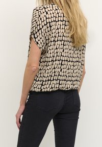 Chemise à manches courtes avec un motif géométrique crème et noir, en tissu léger. Associée à un jean foncé ajusté, présentant un look décontracté.