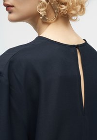 Femme blonde aux cheveux bouclés portant de grandes boucles d'oreilles texturées en forme d'anneau et un haut noir avec une encolure fendue à l'arrière, fermée par un petit bouton.