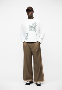 Άνδρας που φοράει άσπρο γραφικό φούτερ, φαρδιά καφέ παντελόνια, ασπρόμαυρα παπούτσια και σκούφο σκούρου καφέ, στέκεται με τα χέρια στις τσέπες.