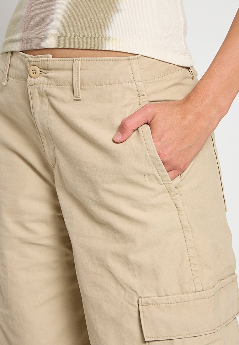 Pantalones cortos de carga beige fabricados con una mezcla de algodón, con bolsillos laterales y cierre de botón. La tela tiene una textura suave y un ajuste relajado.