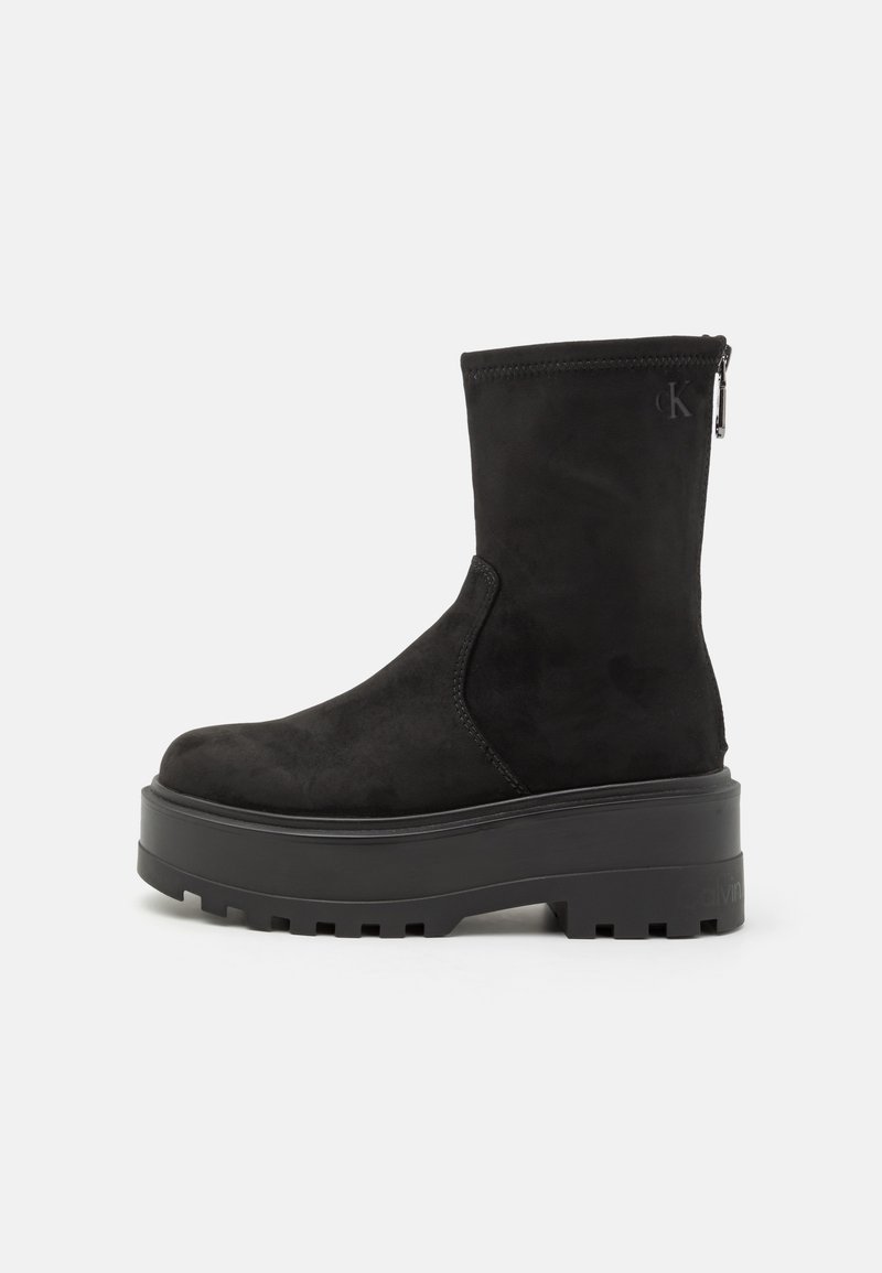 Bota de ante negro hasta la mitad de la pantorrilla con suela gruesa de plataforma, suela exterior acanalada, cremallera en la parte trasera y sutil logo "cK" cerca de la parte superior.