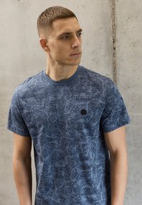 Modré tričko s krátkými rukávy, zdobené texturovaným vzorem v tmavší modré, kulatý výstřih a malým černým logem na levé straně hrudi.
