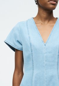 Γυναίκα που φοράει ανοιχτό γαλάζιο τοπ από τζιν με κοντά μανίκια, λαιμόκοψη σε σχήμα V και κάθετη ραφή, φαίνεται από τους ώμους έως το μέσο του κορμού.