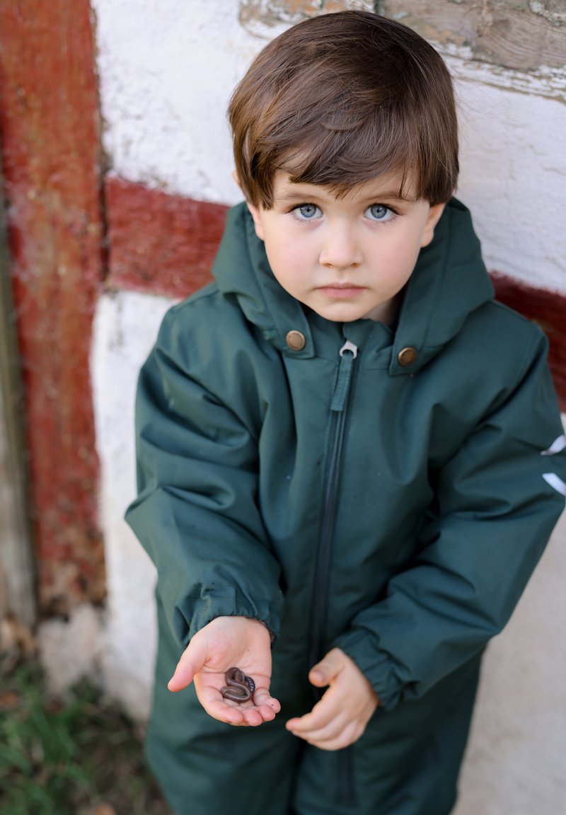 Cappotto impermeabile verde scuro, con zip e bottoni a scatto. La mano di un bambino tiene diversi vermi marroni. Lo sfondo presenta muri rustici rossi e bianchi.