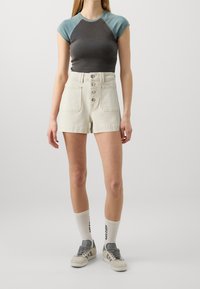 Top corto en gris y verde azulado con cuello redondo, combinado con pantalones cortos de tiro alto en beige que presentan detalles de botones y dos bolsillos frontales.