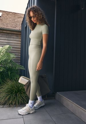 Svetlo zelena športna oblačila, ki vključujejo oprijet zgornji del s kratkimi rokavi in visokim pasom legginsov. Kombinirano s kremnimi supergami in rjavo torbo za na ramo.