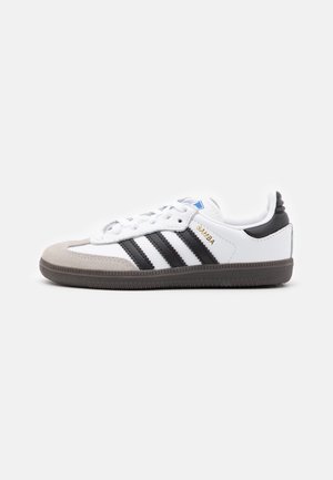 Hvid lav Adidas Samba sneaker med sorte striber, grå ruskindstot, sort hælflap og brun sål, vist fra siden.