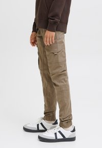 Beżowe spodnie typu cargo z wieloma kieszeniami, elastycznymi mankietami i prostym krojem nogawek, zestawione z białymi sneakersami z czarnymi akcentami.