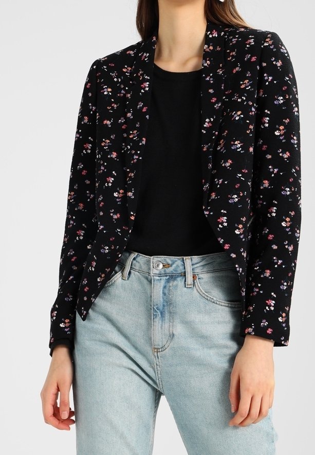 Blazer noir à motif floral avec une coupe ajustée, présentant de petites fleurs colorées, un col à revers et des manches longues, porté sur un haut noir et un jean clair.